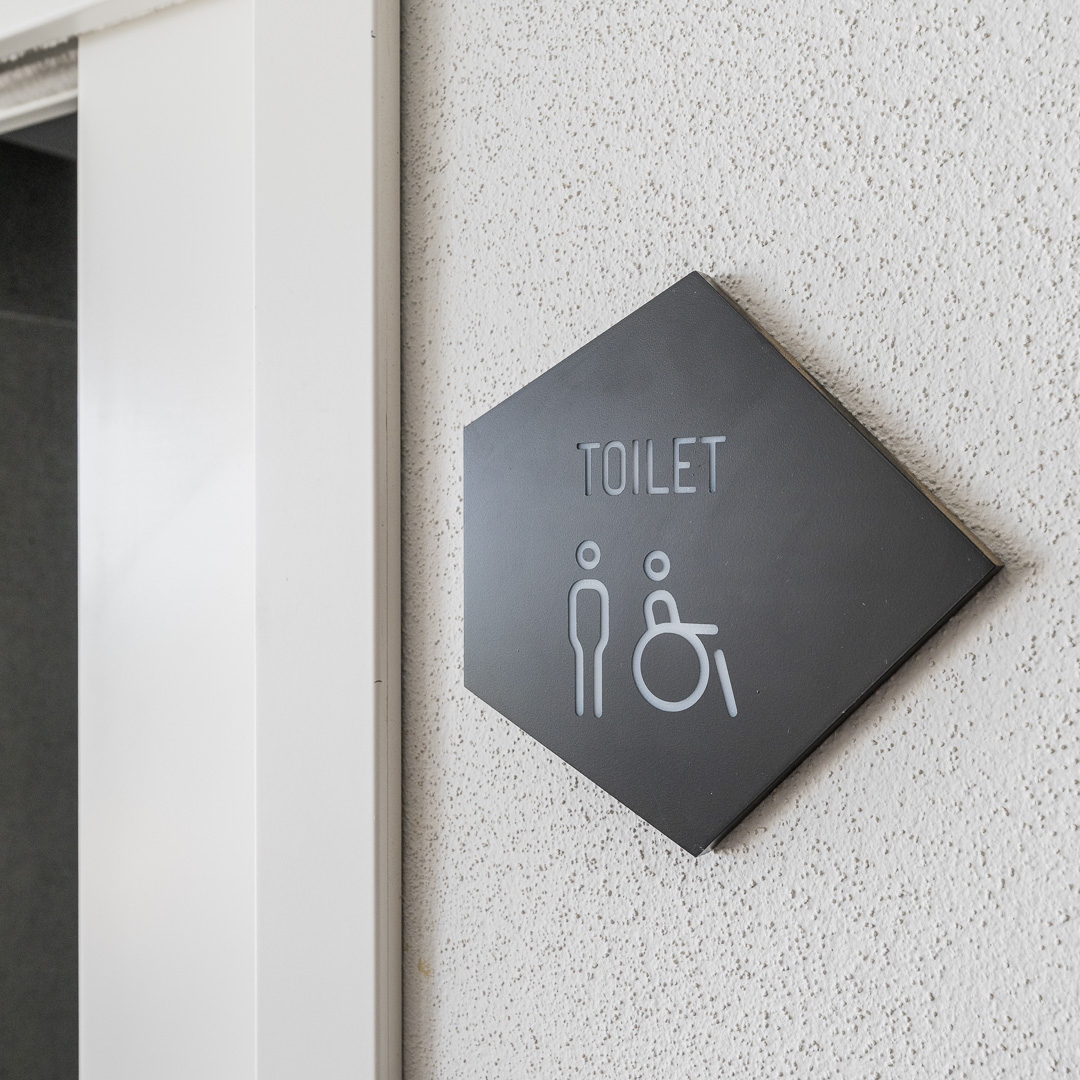 Placa negra con la palabra “Toilet” que indica el baño, colocada sobre una pared blanca en una zona común.