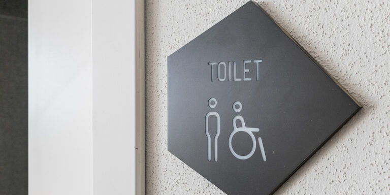 Placa negra con la palabra “Toilet” que indica el baño, colocada sobre una pared blanca en una zona común.