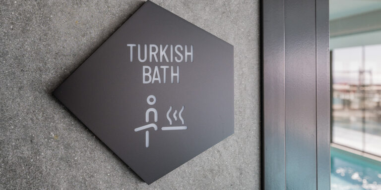 Placa negra con la palabra “Bath Turkish” que indica el baño turco, colocada sobre una pared blanca en una zona común.