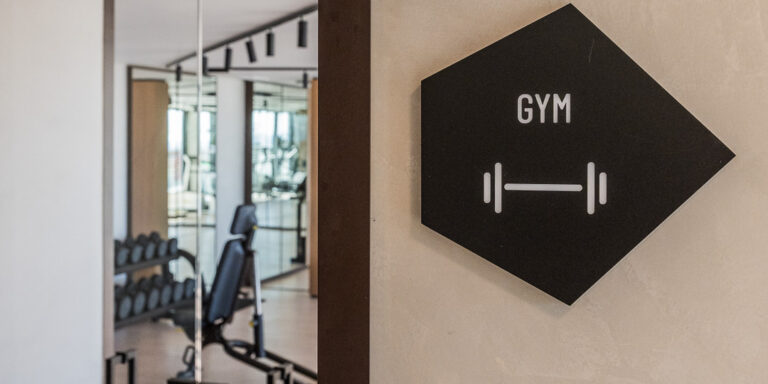 Placa negra con la palabra “Gym” que indica la presencia de un gimnasio, colocada en una pared blanca.
